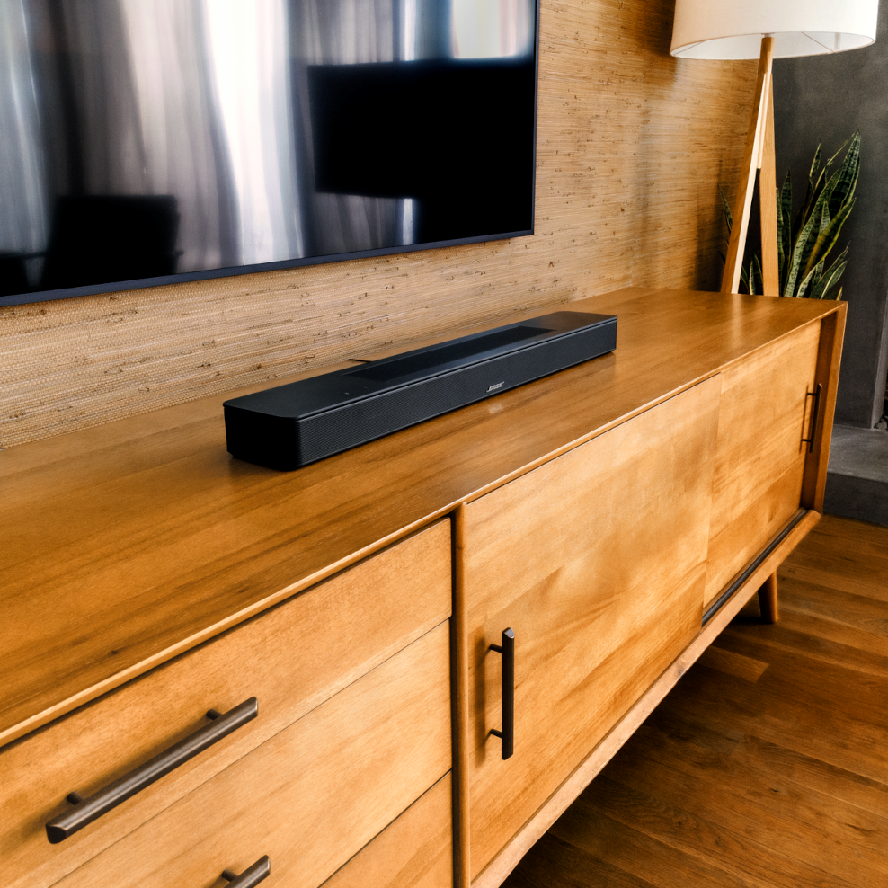 Een zwarte soundbar op een houten kast onder een televisie. Een vloerlamp staat op de achtergrond rechts.