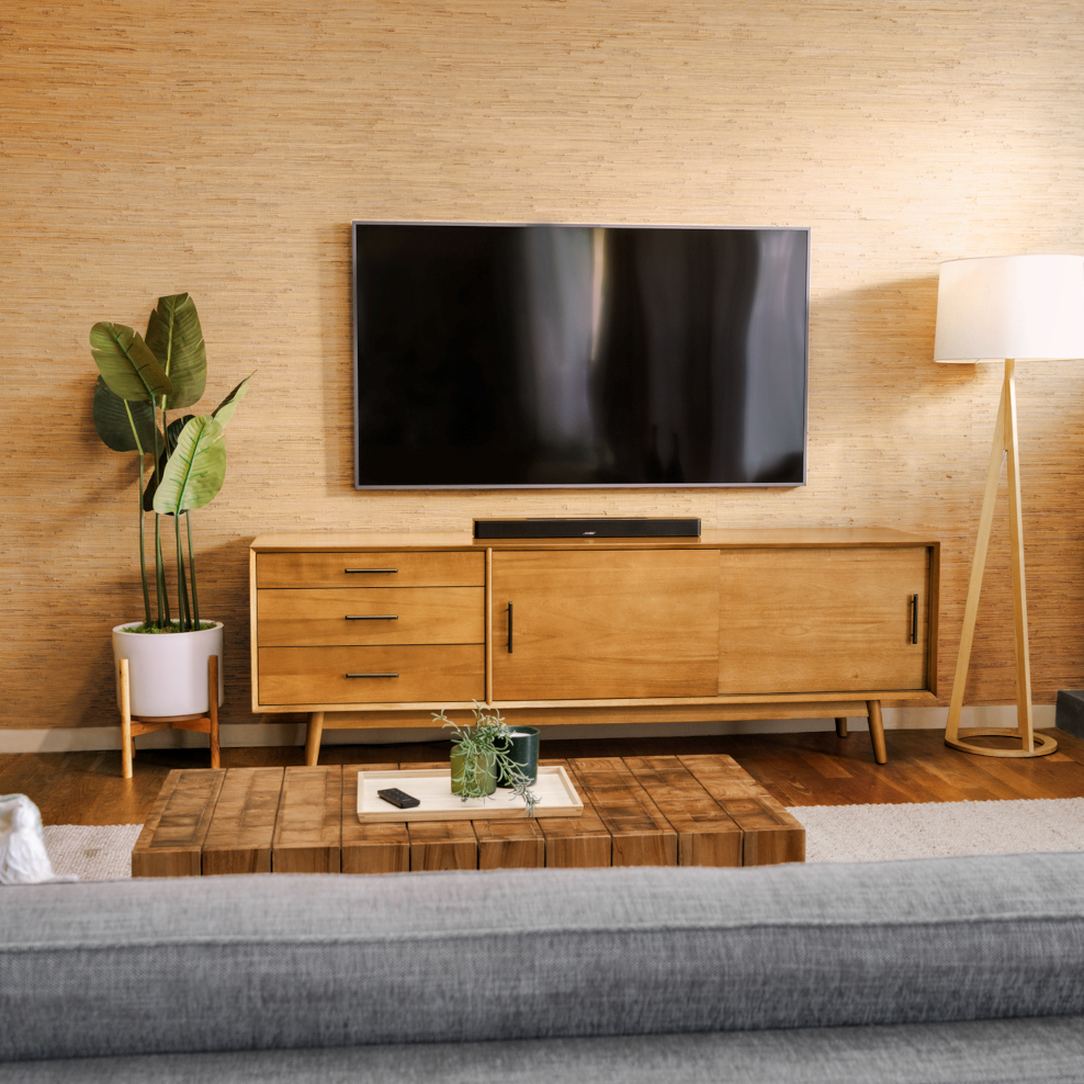 Woonkamer met houten tv-meubel, tv, plant en een grijze bank. Houten vloer en een lichtgekleurde muur.