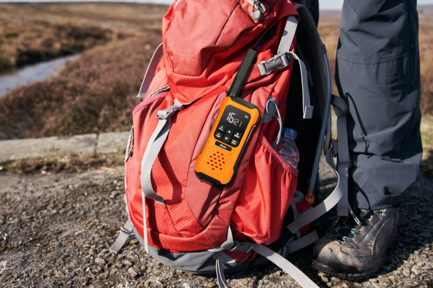 A backpack with a walkie-talkie, water bottle, on it. The person is wearing hiking boots and pants.