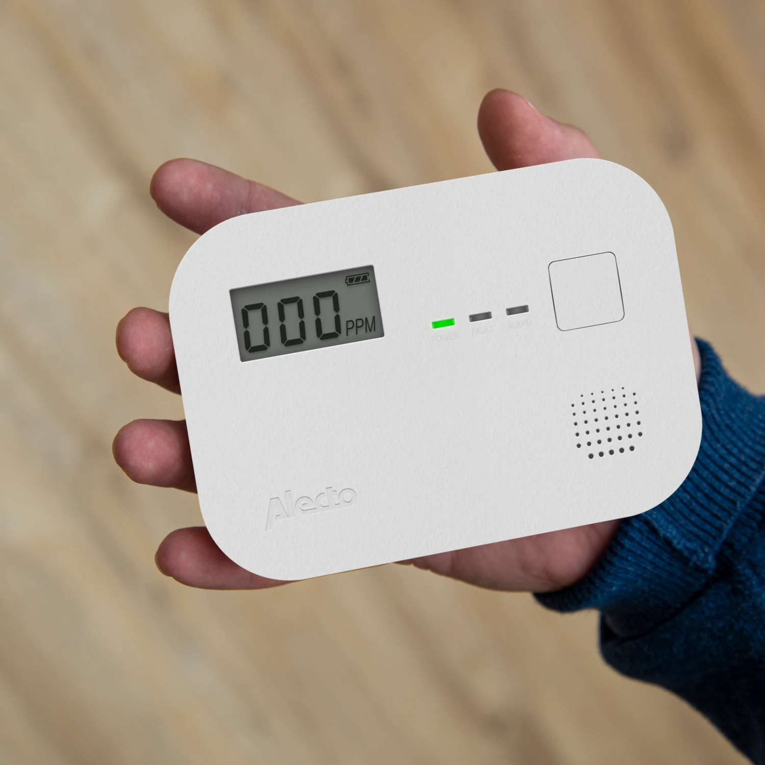A person's hand holding a white carbon monoxide detector. Display shows 000 PPM. Green light.