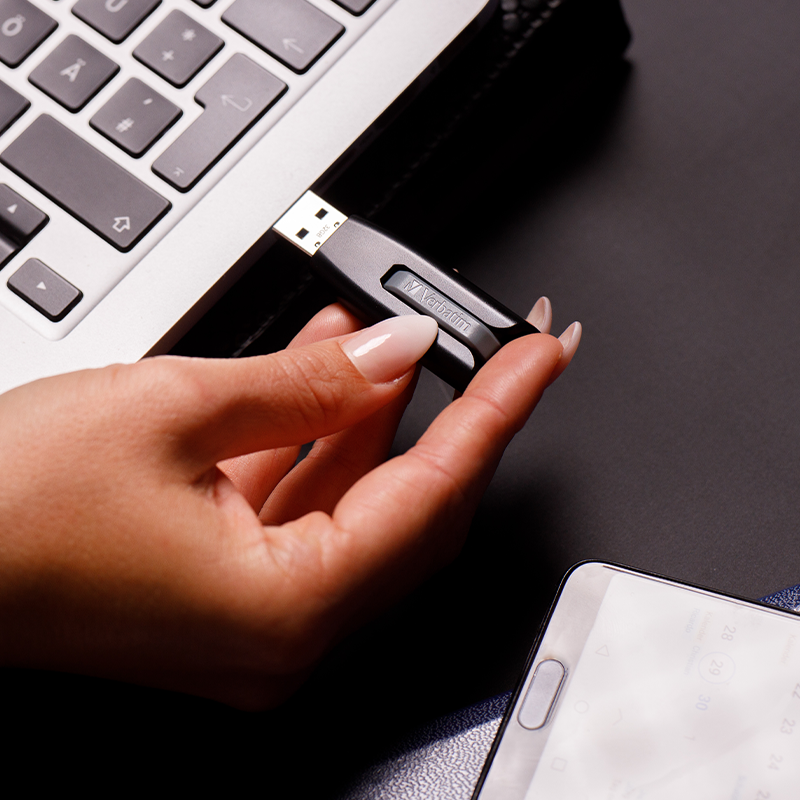 El, siyah bir USB sürücüyü dizüstü bilgisayara takıyor, bir akıllı telefon görünüyor.