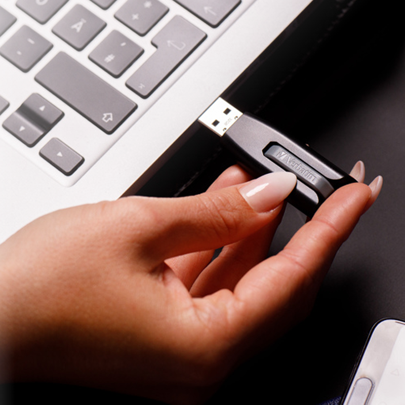 Bir el, siyah bir USB sürücüyü bir dizüstü bilgisayara takıyor. Sürücüde Verbatim logosu var.