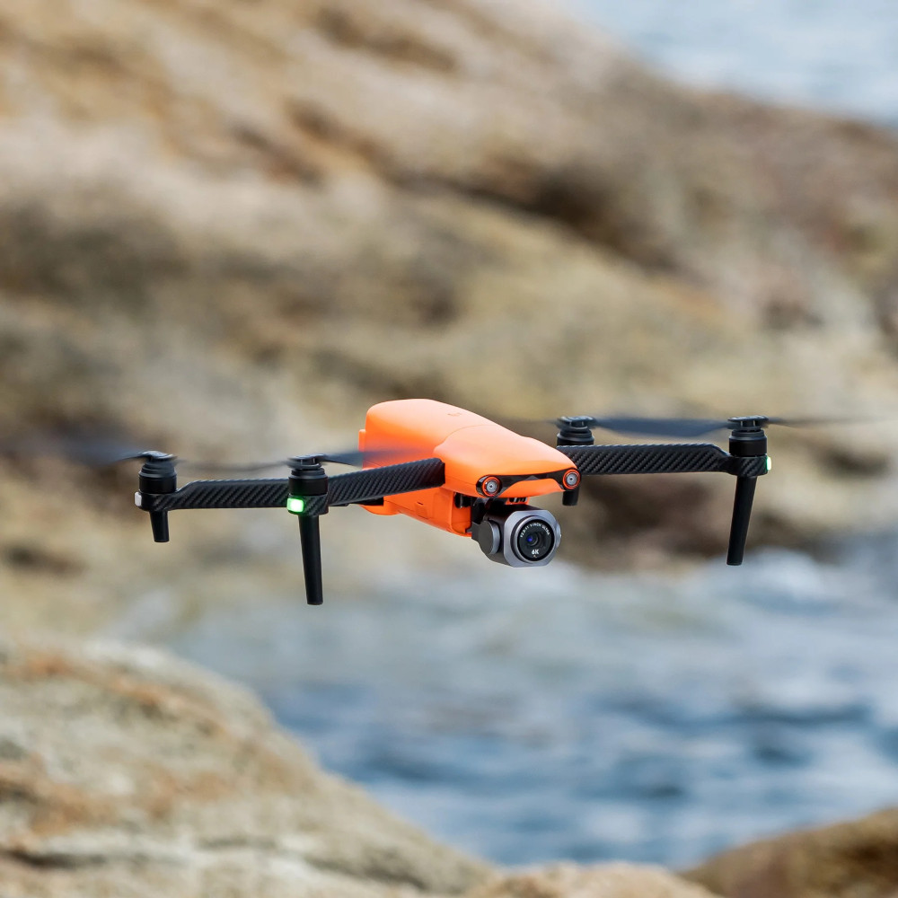 An orange drone in flight. The background shows rocks and water.
