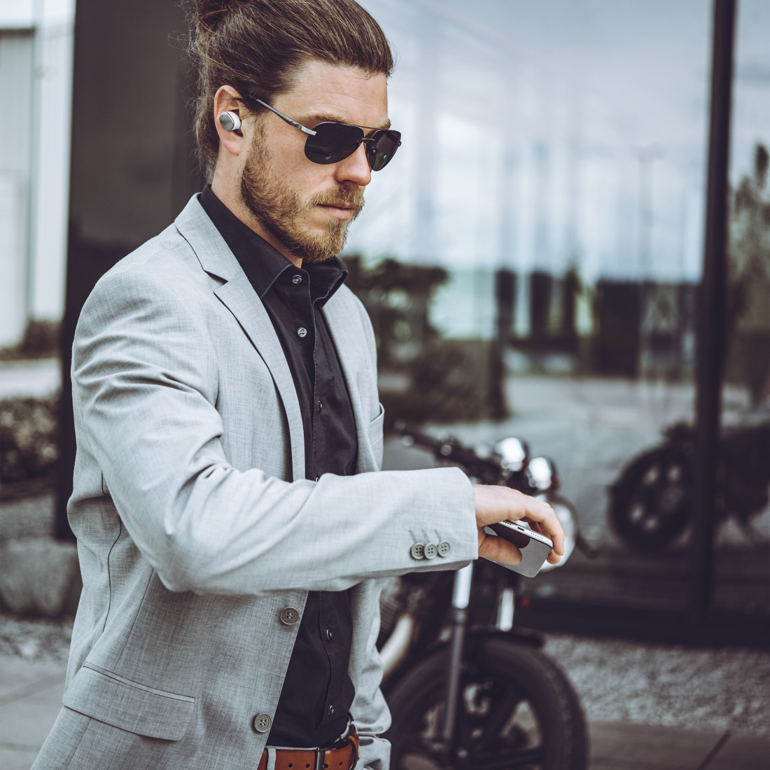 Mann mit Sonnenbrille und Blazer, trägt Ohrhörer, hält Telefon. Motorrad im Hintergrund.