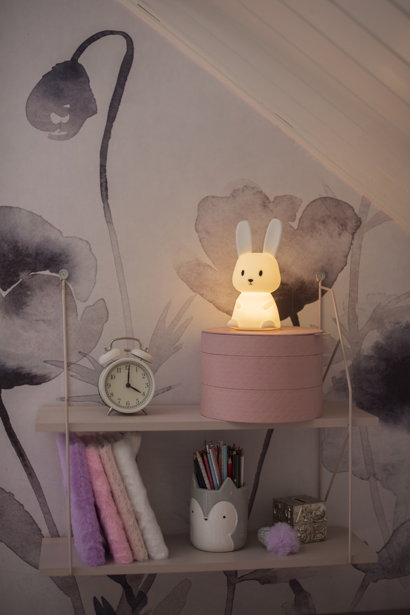 Una lampada a forma di coniglietto illuminata su una mensola, con un orologio, matite e decorazioni.