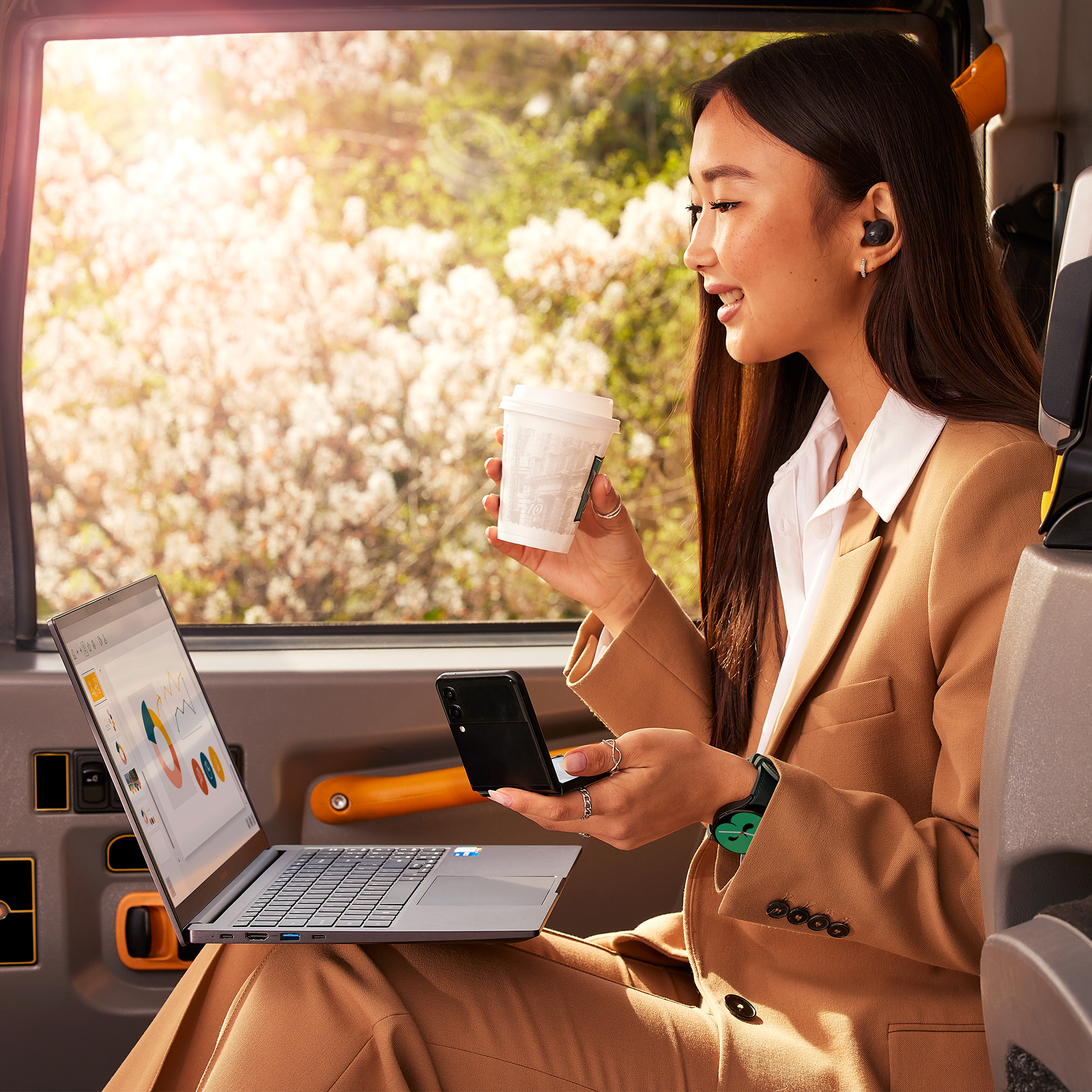 Mujer en traje en autobús, usa portátil, sostiene café, escucha música con auriculares.