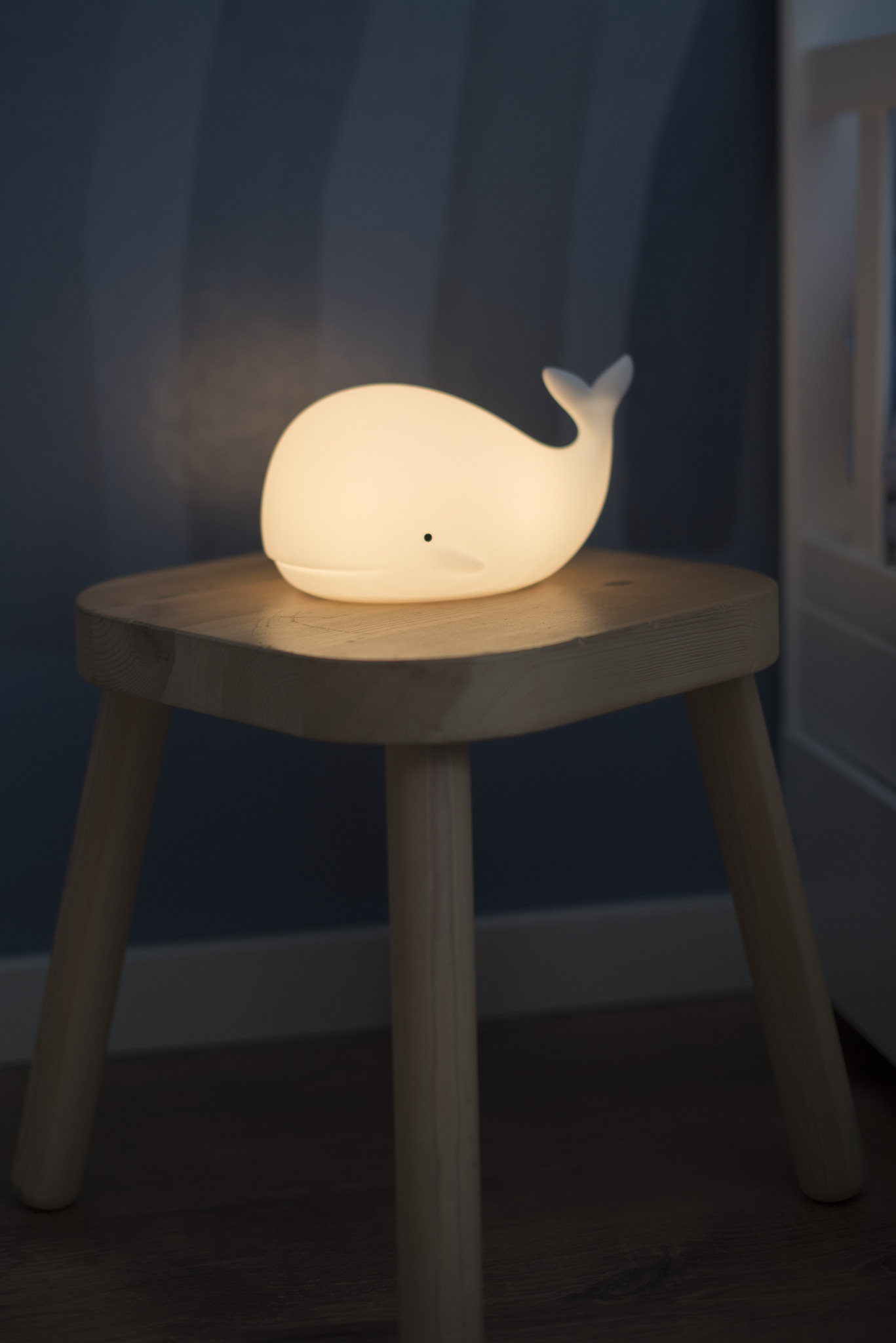 Una luce notturna a forma di balena su uno sgabello di legno. Emette luce soffusa.