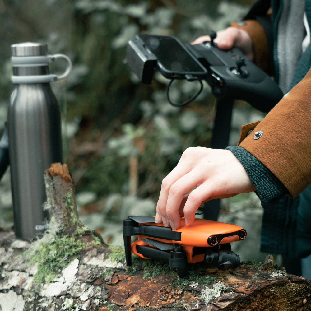 Handen van een persoon die een drone bedient met een controller en telefoon, op een boomstam, in de natuur.
