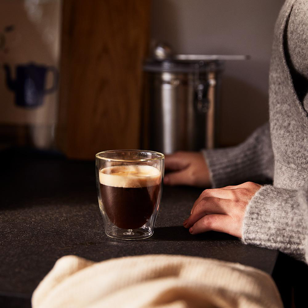 Ein Glas mit Kaffee, auf einem dunklen Tisch. Hände und Arme einer Person sind im Hintergrund sichtbar.