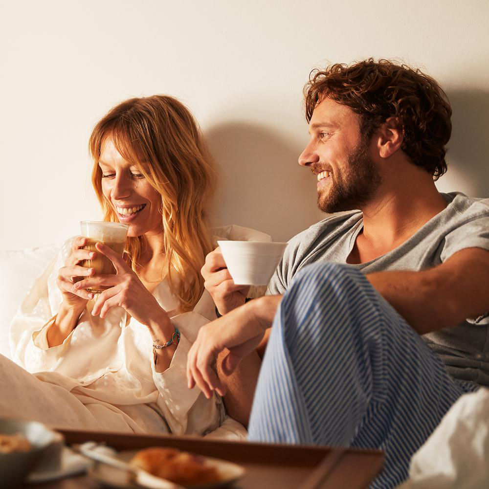 Paar im Bett, trinkt Kaffee, lächelt. Tablett mit Essen auf dem Bett.