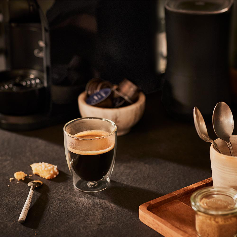 Kaffeeglas auf schwarzem Tisch mit Kaffeepads, Kaffeemaschine und Zucker.
