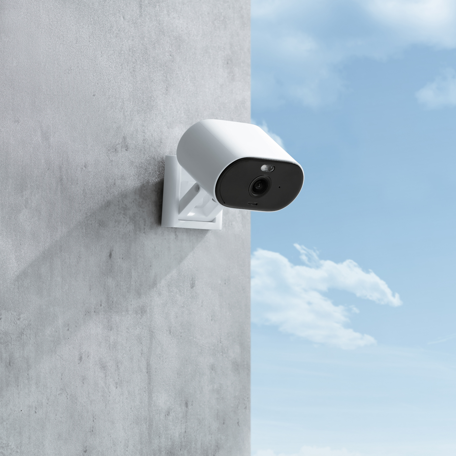 Caméra de sécurité blanche montée sur un mur gris, ciel bleu avec des nuages.