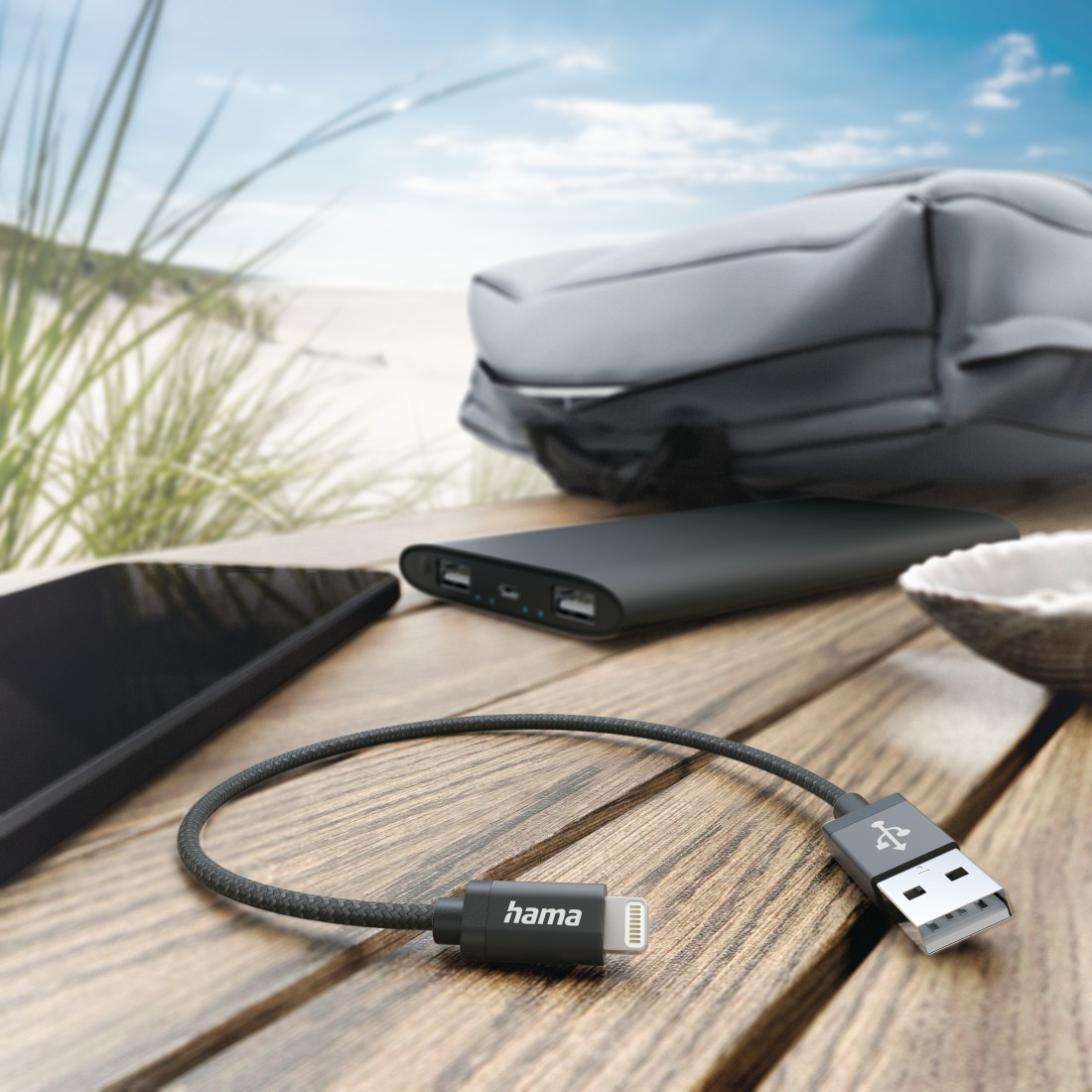 Ein USB-Kabel mit Telefon, Powerbank und Tasche auf einem Holztisch, Strand im Hintergrund.