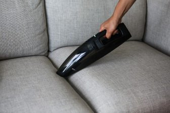 Hand vacuuming a gray sofa with a black handheld vacuum cleaner.