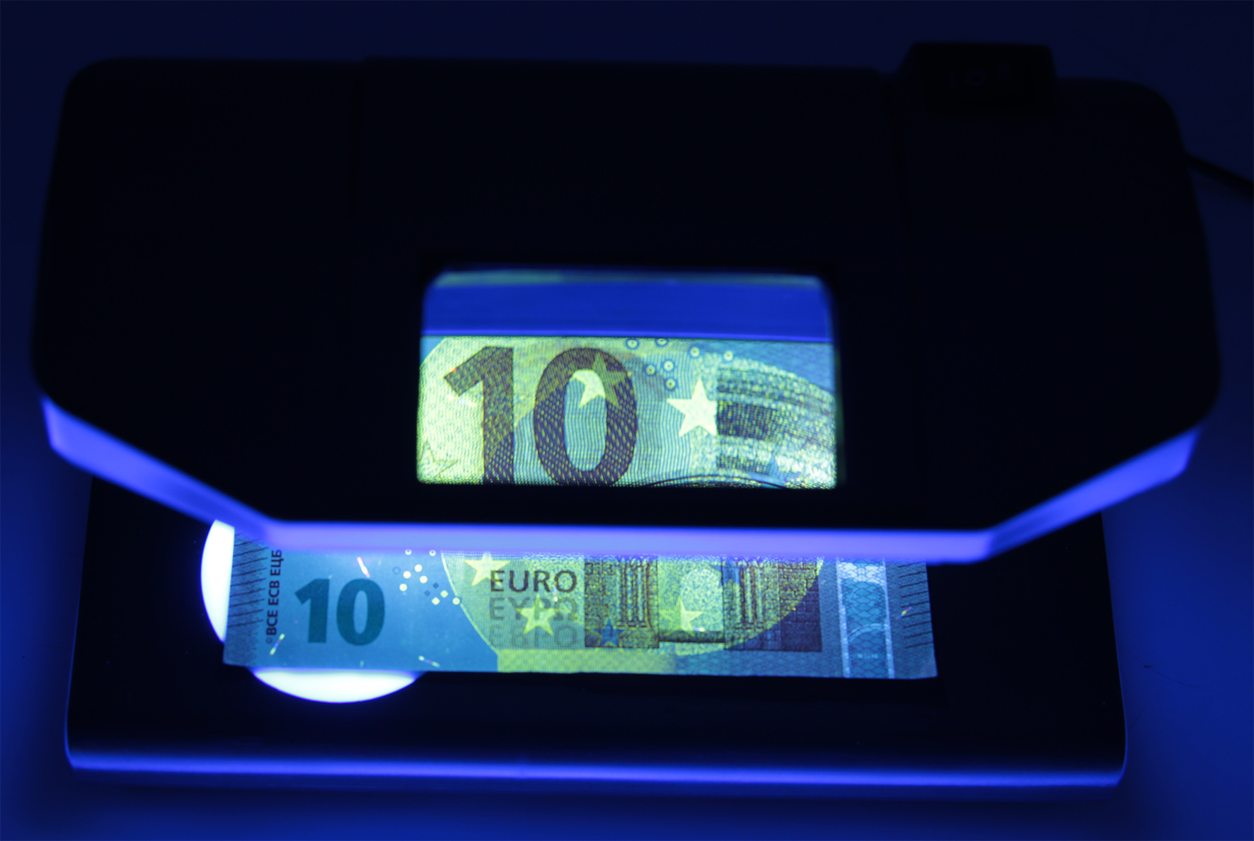 Egy fekete eszköz 10 eurós bankjegyet világít meg UV-fénnyel, felfedve a biztonsági funkciókat.