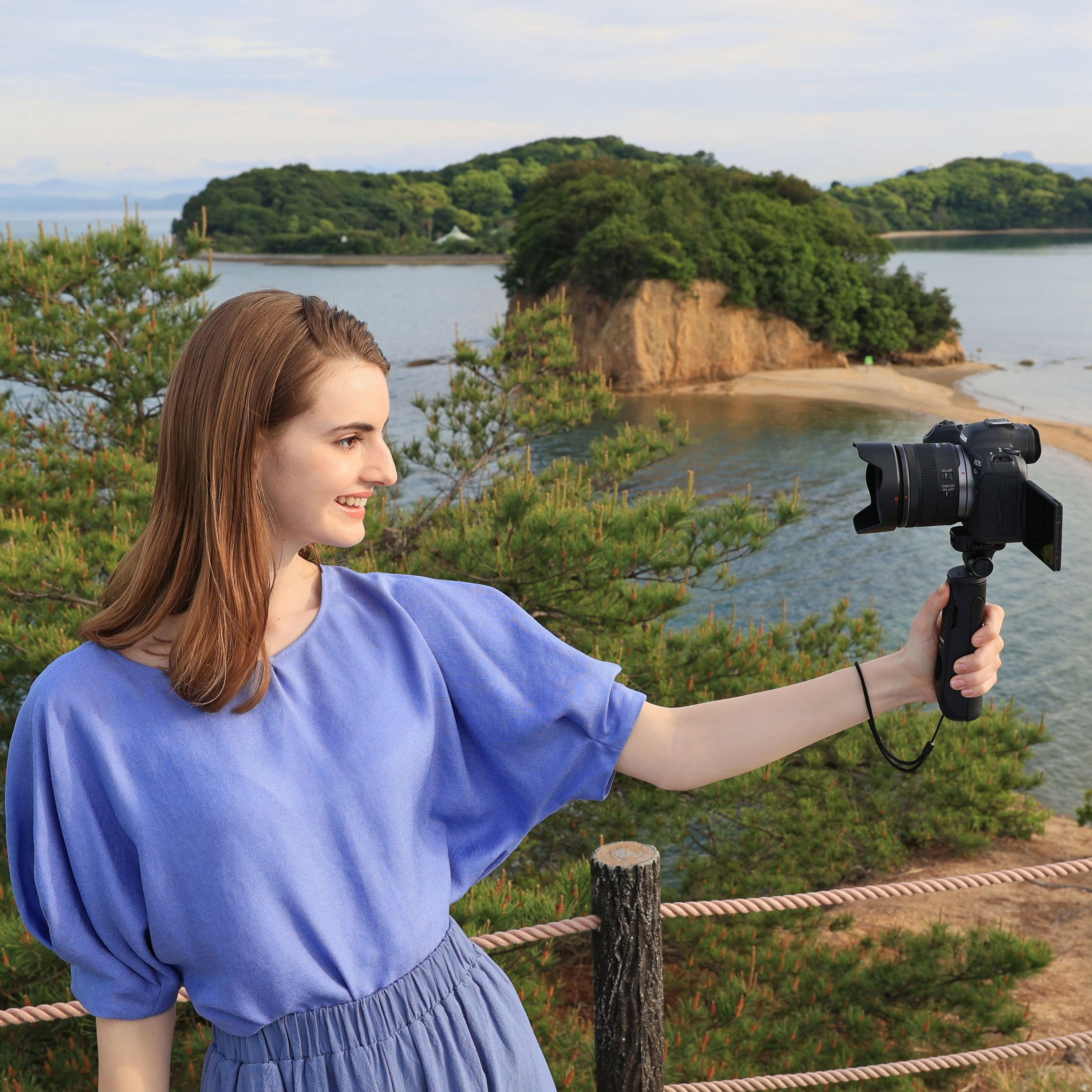 Una donna in top blu si riprende con una telecamera su impugnatura davanti a un paesaggio costiero.