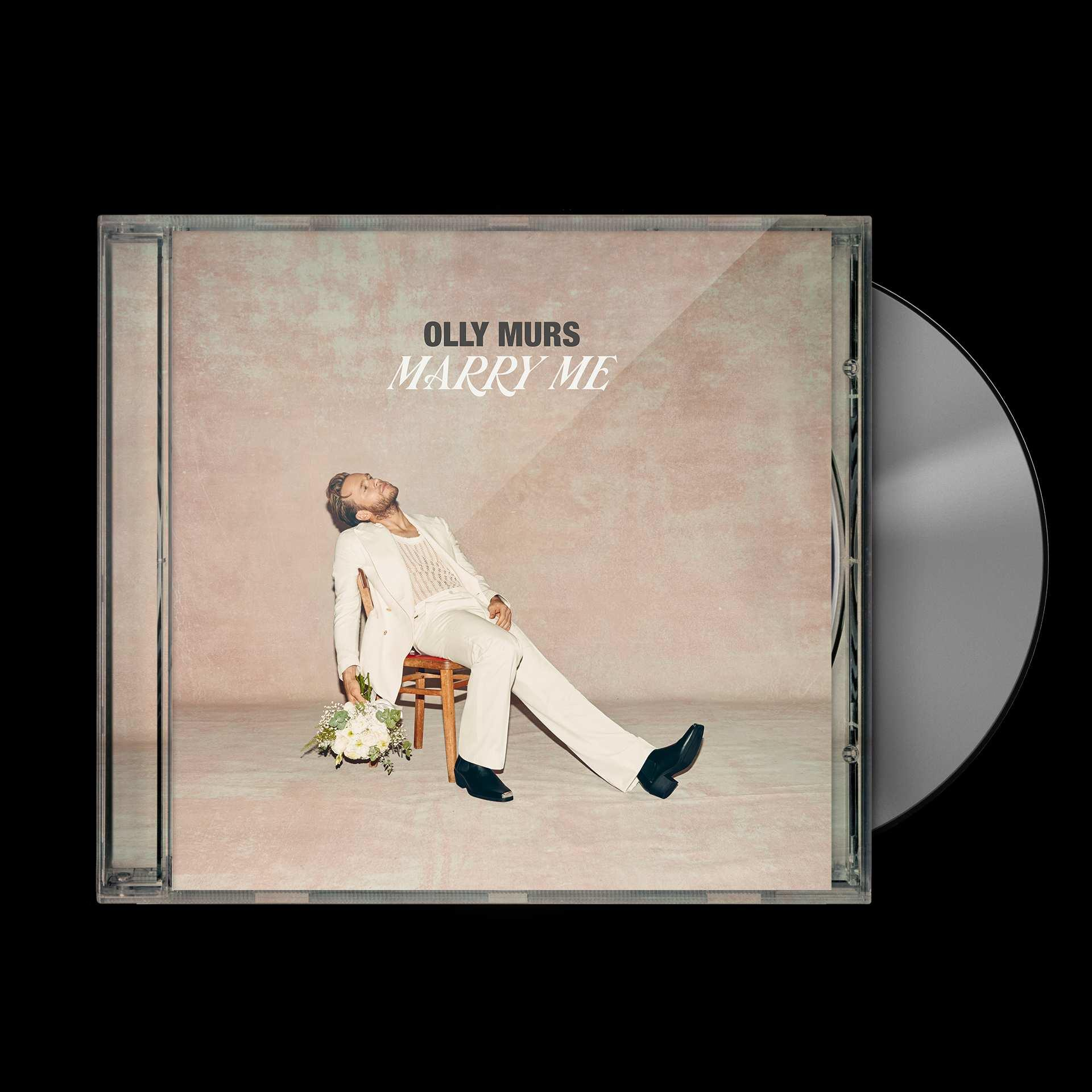 Albumcover von Olly Murs' 'Marry Me'. Mann im weißen Anzug auf einem Stuhl mit Blumen, vor pinkfarbenem Hintergrund.