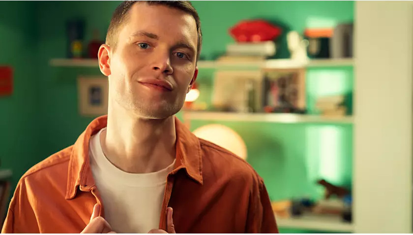 Un homme en chemise orange et t-shirt blanc, souriant. Il est devant un mur vert.