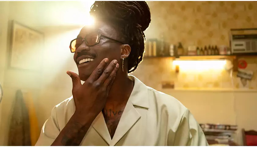 Un homme avec une moustache se touche le visage. Il porte des lunettes et une chemise claire. Le fond montre une pièce.