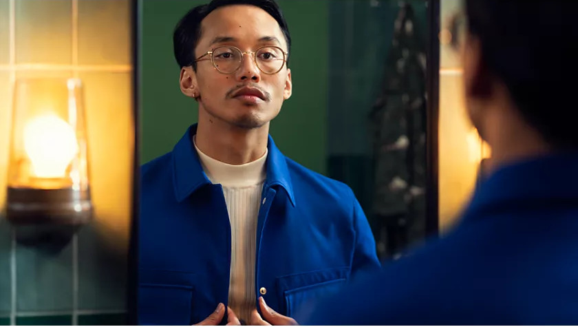 Homme en veste bleue se regardant dans un miroir. Il porte des lunettes et une moustache.