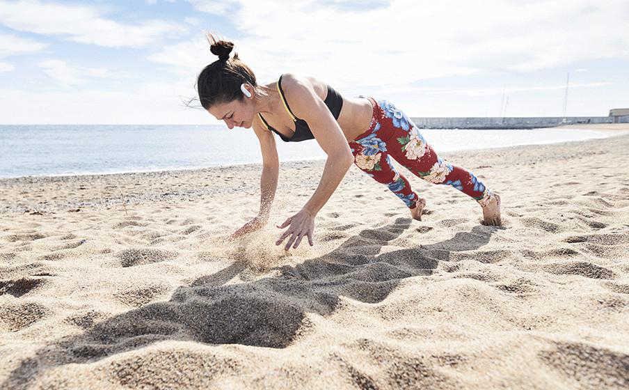 Frau macht Liegestütze am Strand. Sie trägt Leggings und ein schwarzes Top. Sand und Meer im Hintergrund.