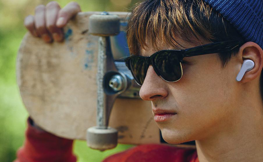 Ein junger Mann mit Sonnenbrille und Ohrhörern, der ein Skateboard hält.