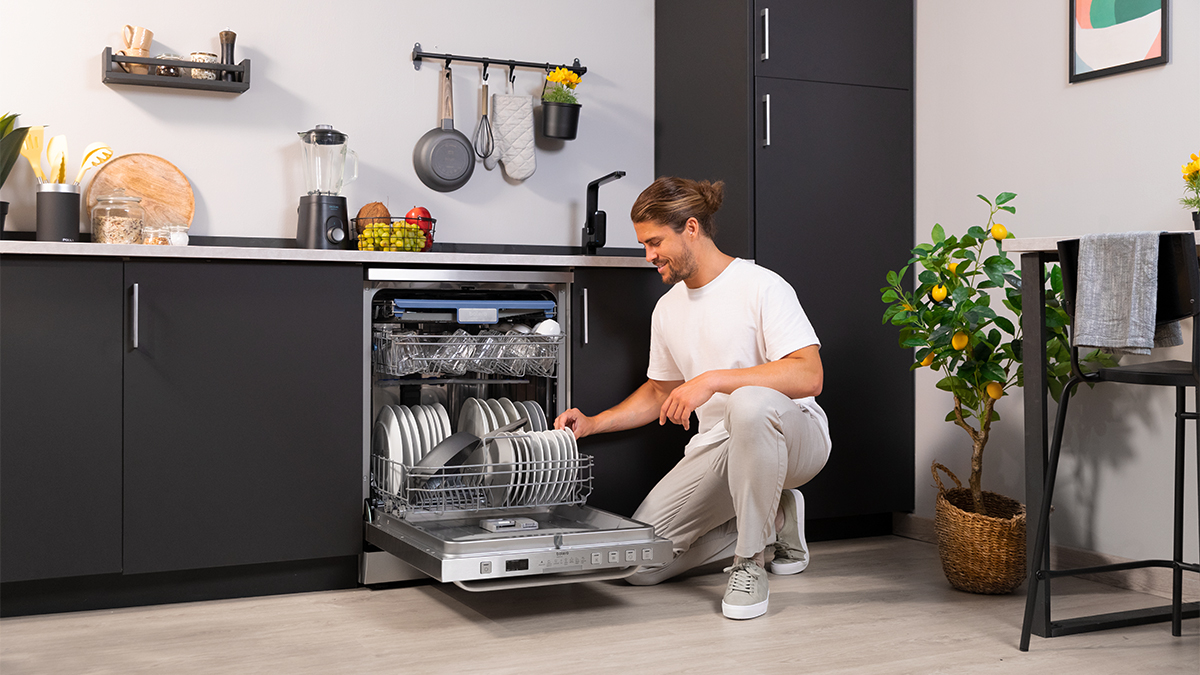 Hombre cargando un lavavajillas con platos en una cocina moderna, sonriendo mientras lo hace.