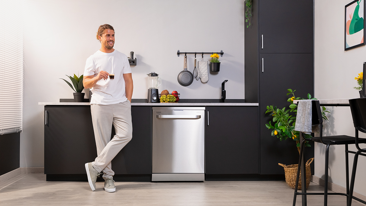 Un hombre en una cocina se apoya en una encimera junto a un lavavajillas, sosteniendo una taza.