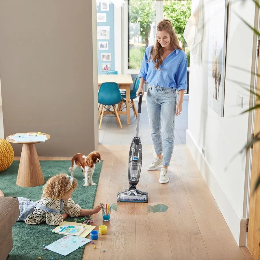 Vrouw stofzuigt een houten vloer terwijl een kind speelt en een hond toekijkt.
