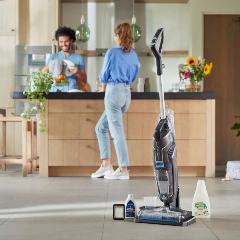 Een schoonmaakscène met een stofzuiger, schoonmaakmiddelen en twee mensen in een keuken.