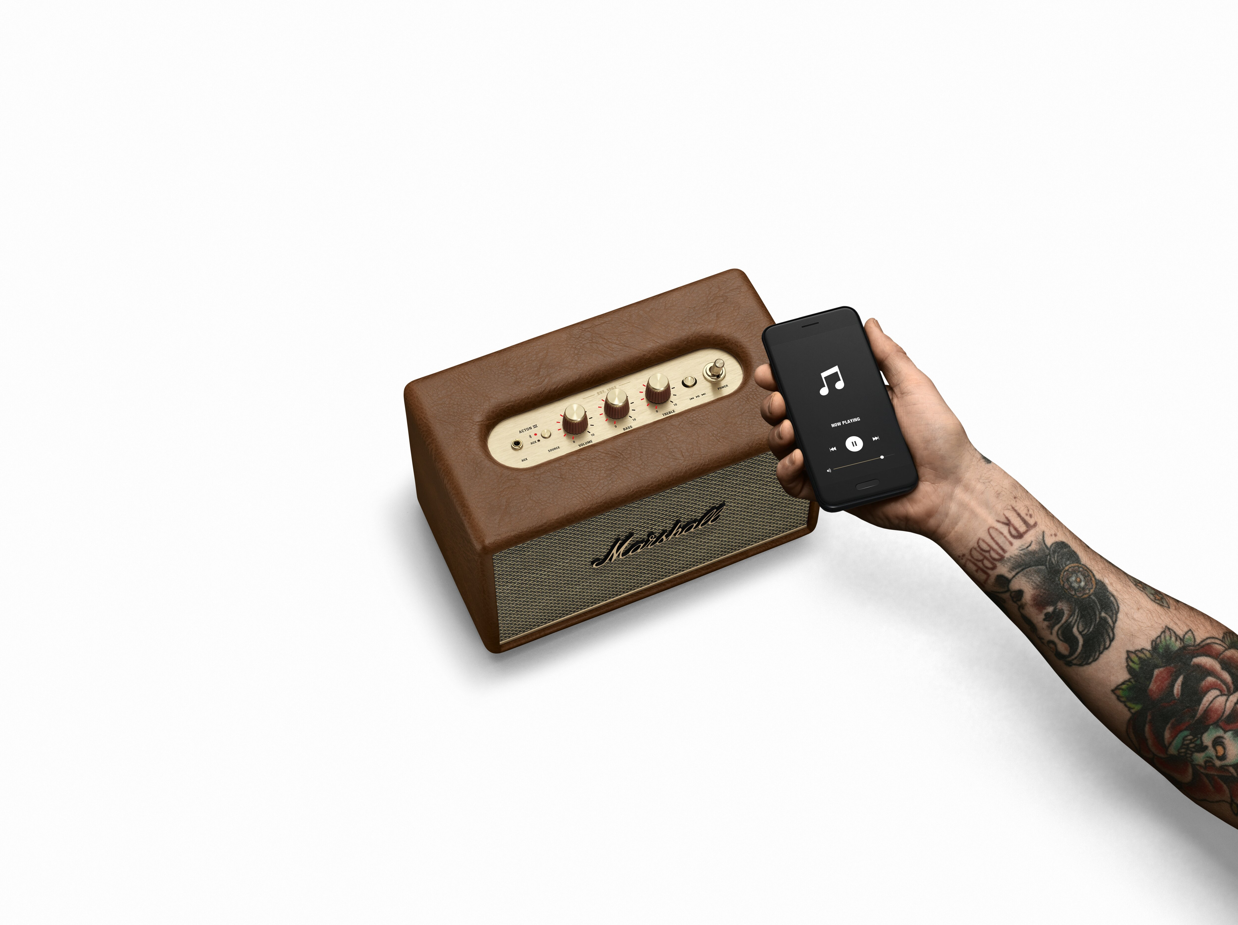 Ein brauner Lautsprecher mit goldenen Knöpfen und ein Handy, das Musik abspielt. Ein Tattoo ist auf dem Arm.