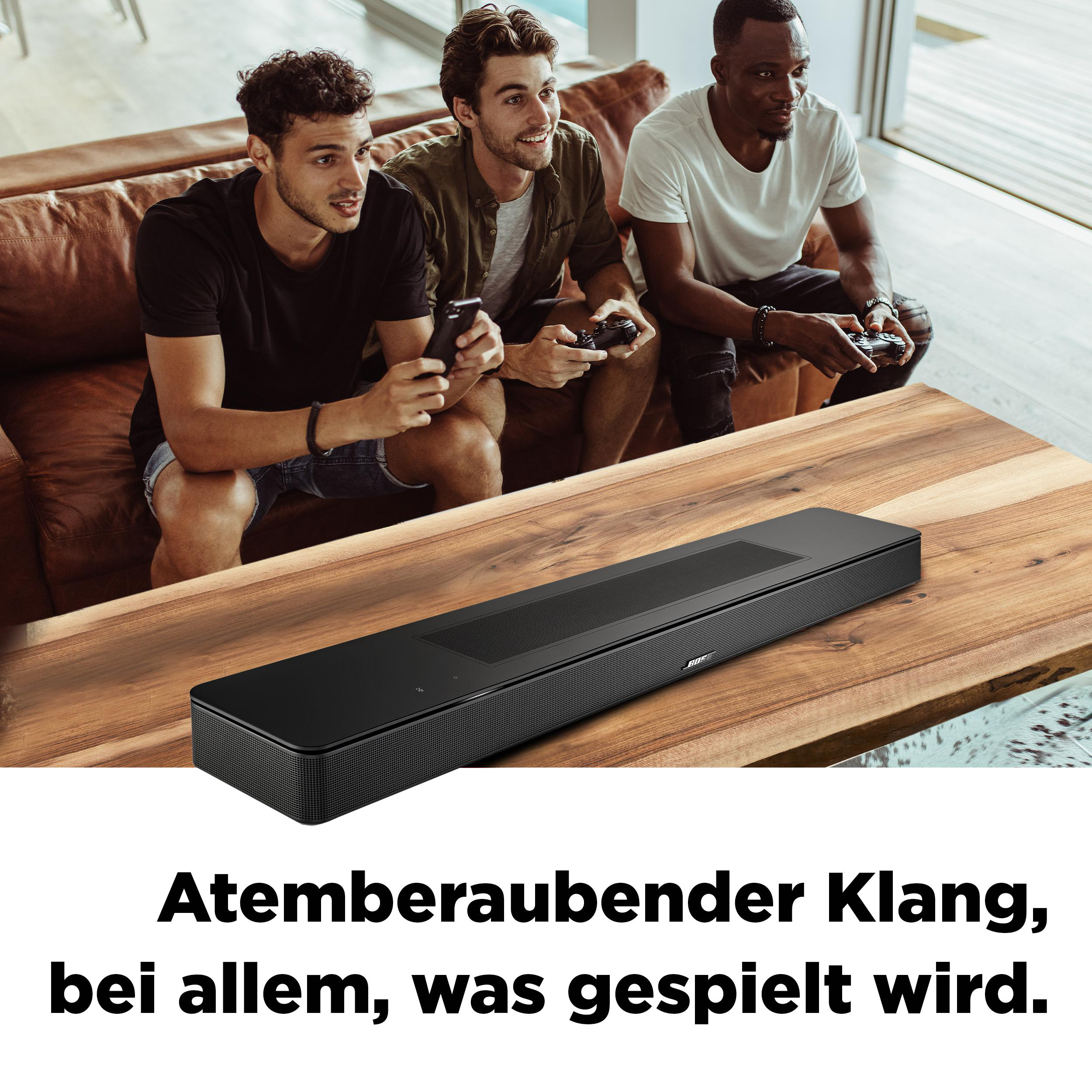 Drei Personen auf einem braunen Sofa spielen Videospiele, mit einer schwarzen Soundbar auf einem Tisch.