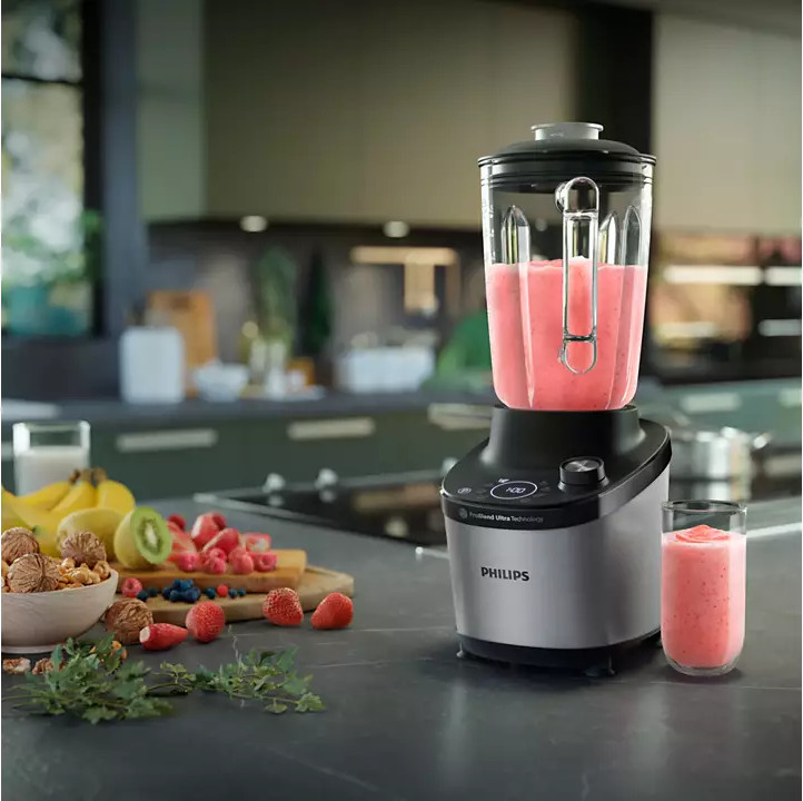 Mixeur avec smoothie rose, et un verre de smoothie, avec des fruits sur la table.