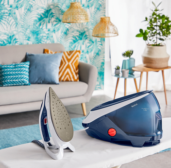 Ein weißes Bügelbrett mit einem Bügeleisen und einem blau-weißen Dampfgenerator. Der Hintergrund ist ein Wohnzimmer.