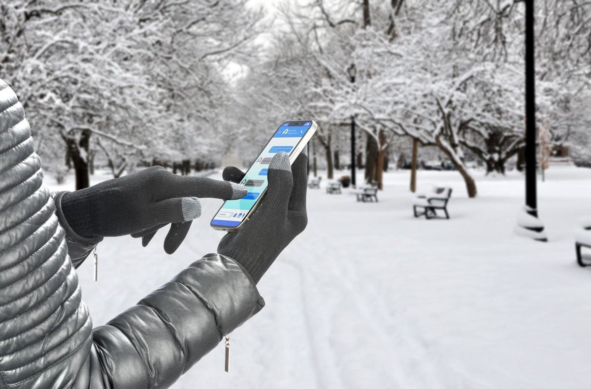 Personne gantée utilisant un téléphone dans un parc enneigé avec bancs et arbres.