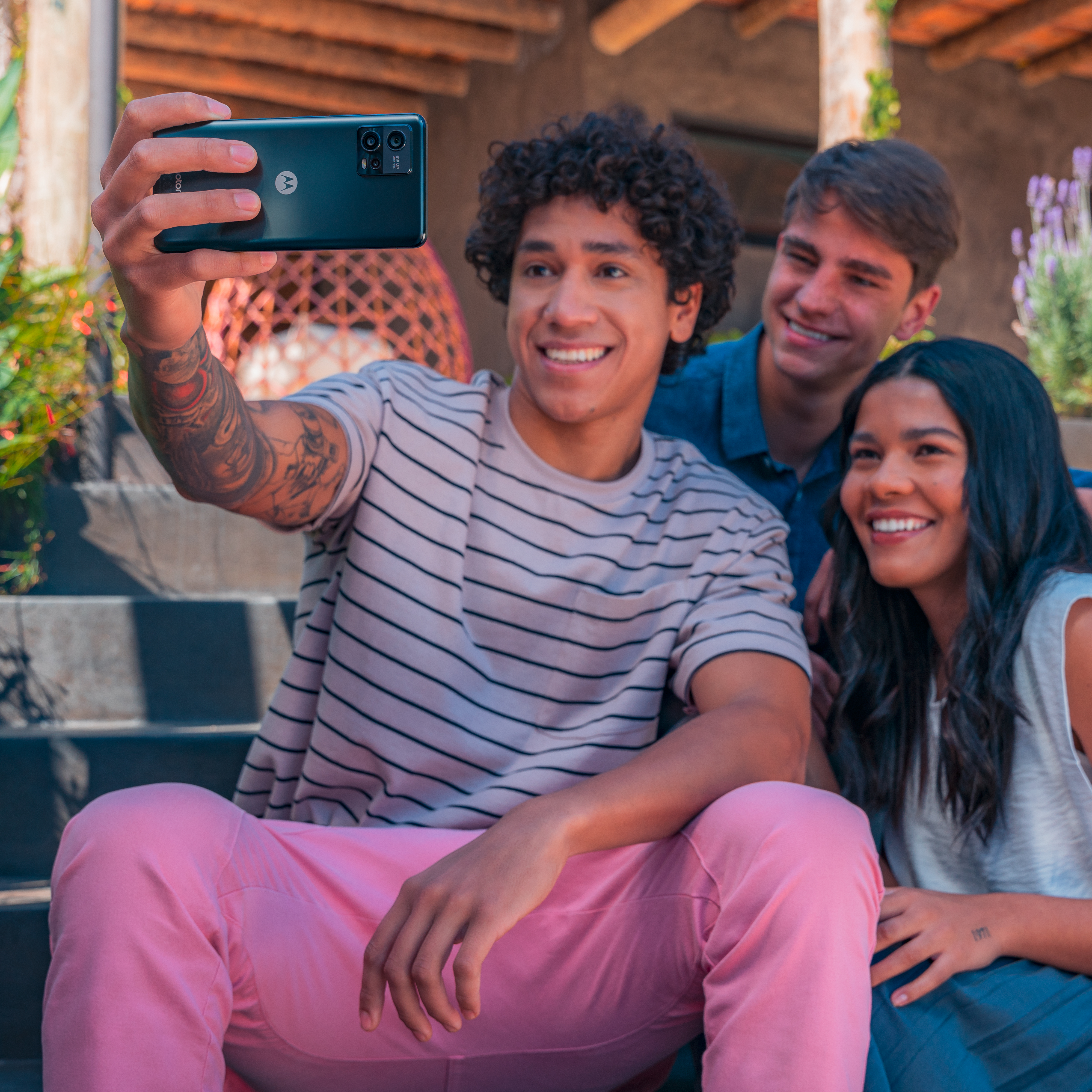 Drei Personen machen ein Selfie. Einer hält das Handy. Im Hintergrund eine sonnige Terrasse.