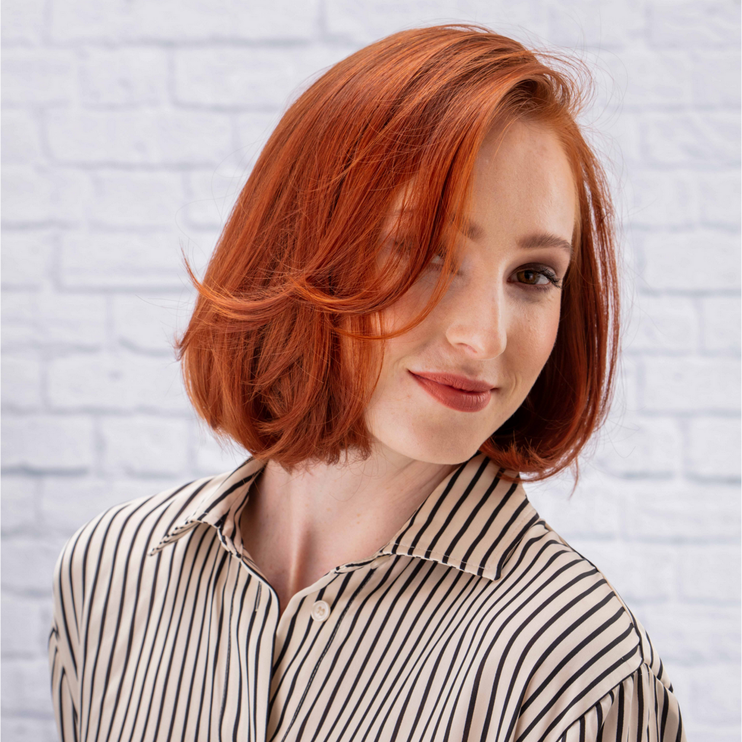 Woman with red hair and striped shirt. White brick wall background, smiling.