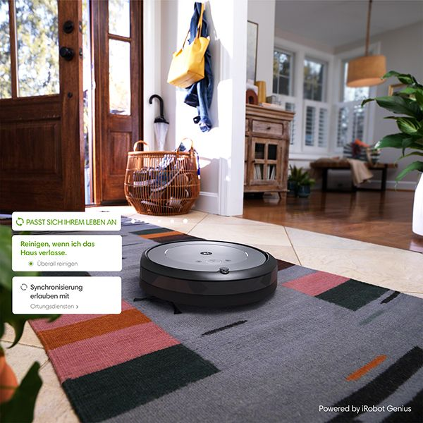 Un aspirateur robot noir et gris sur un tapis à motifs dans un couloir. Une porte est ouverte en arrière-plan.