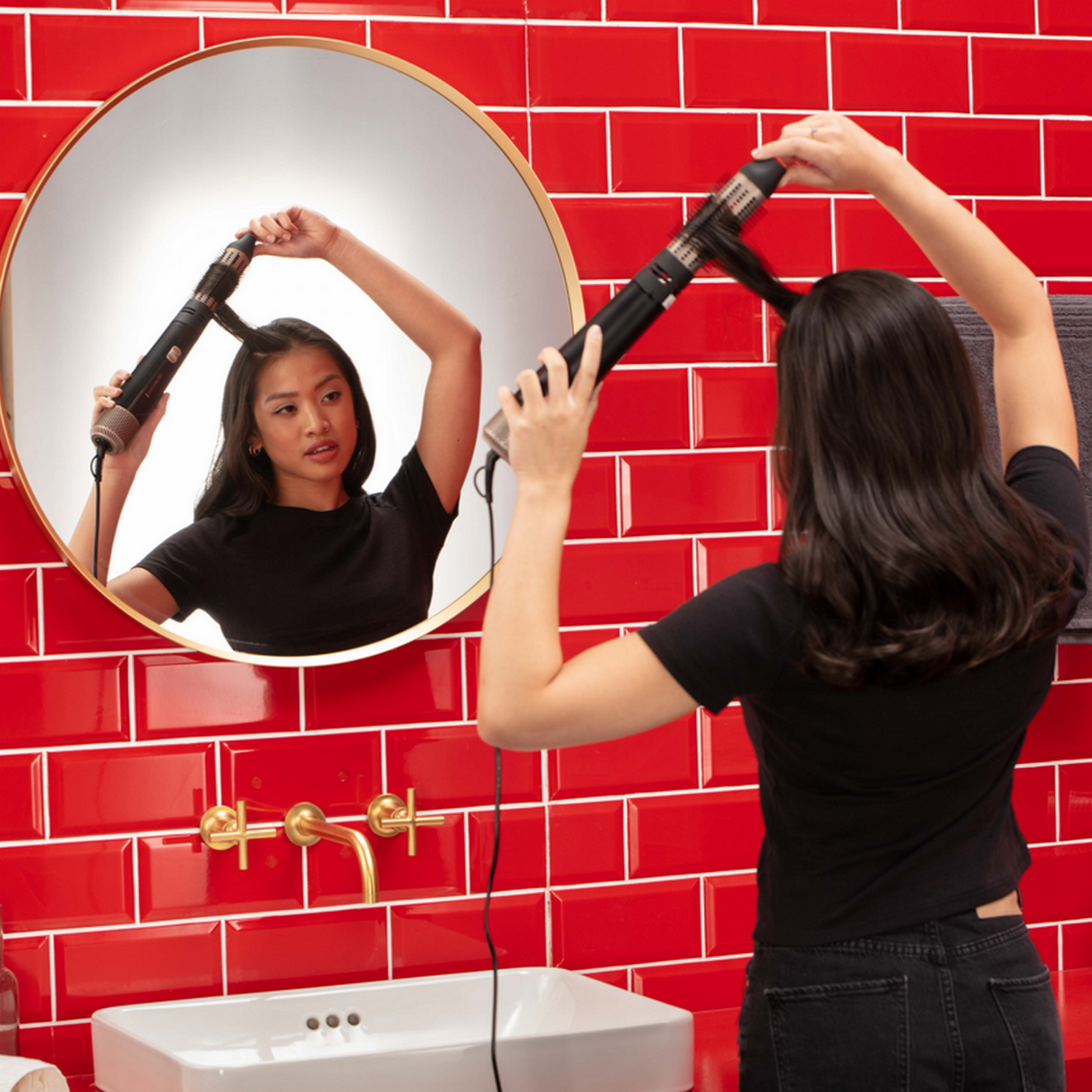 Vrouw die haar haar in de spiegel stylet met een rode tegelachtergrond in een badkamer.