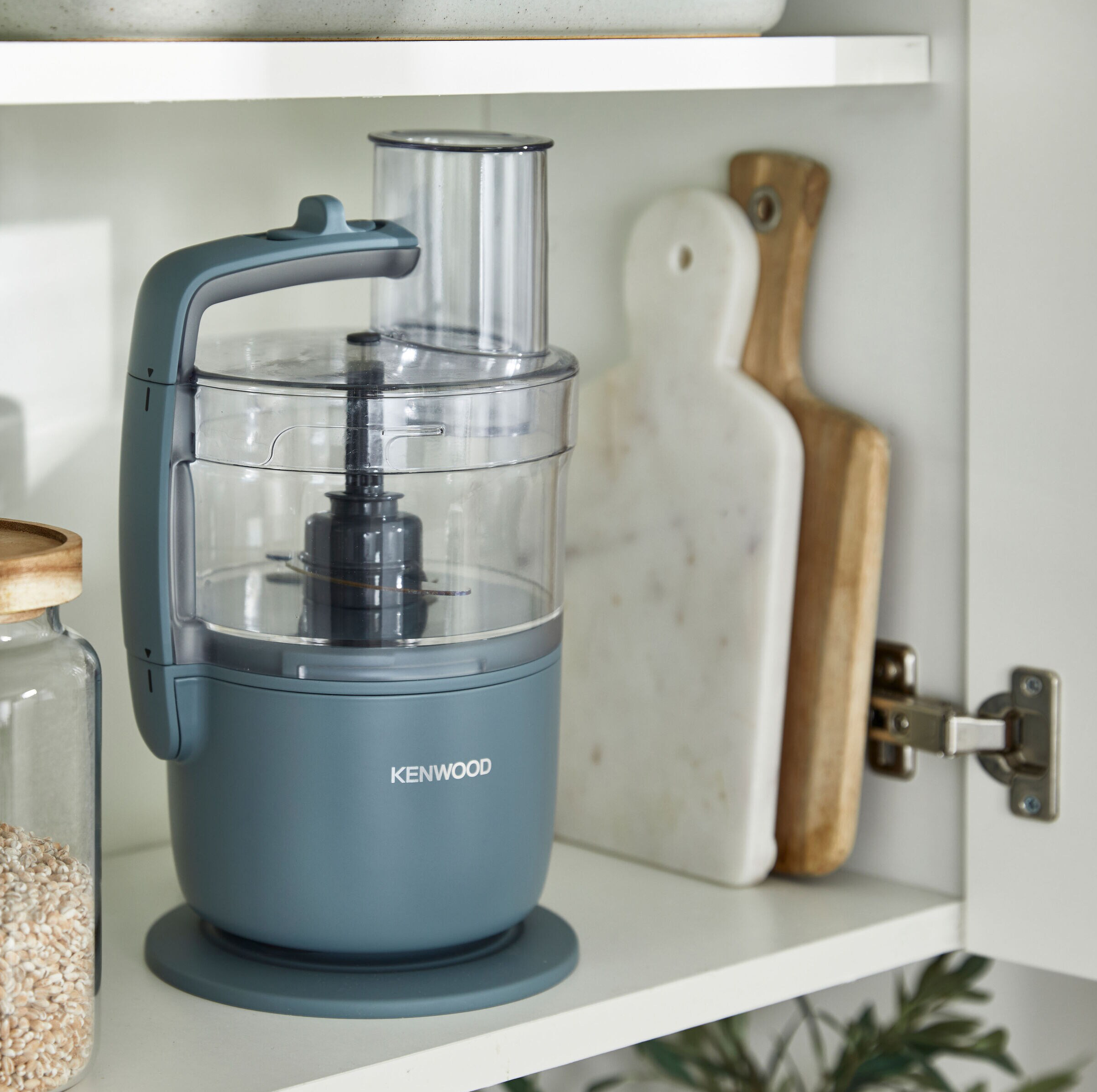 Ein grauer Küchenmixer mit transparentem Behälter, in einem weißen Schrank mit Schneidebrettern.