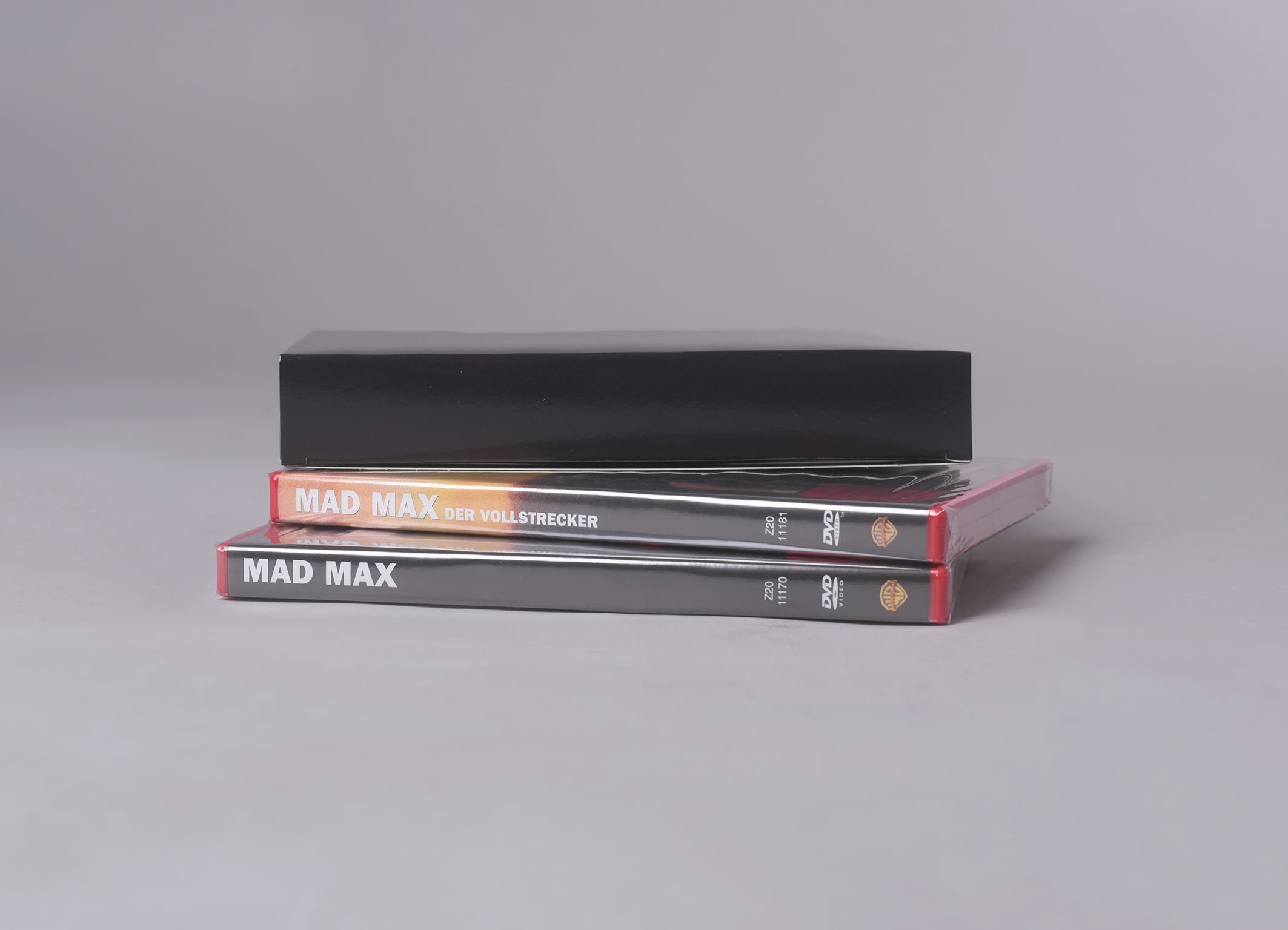 Zwei Mad Max DVDs in Hüllen, gestapelt mit einer schwarzen Box oben, auf grauer Fläche.