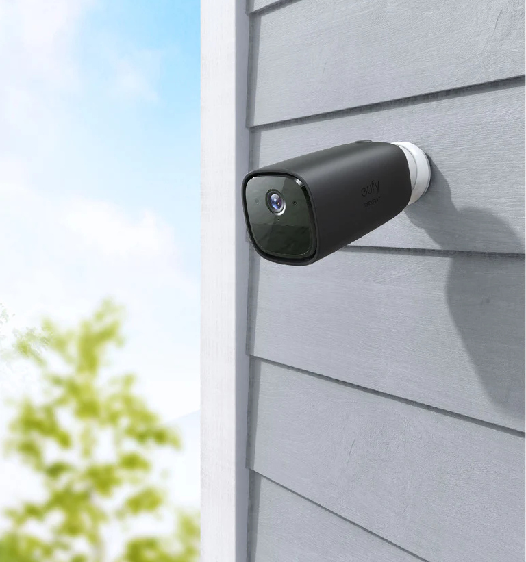 Caméra de sécurité noire fixée sur un mur gris, partiellement cachée par le feuillage vert.