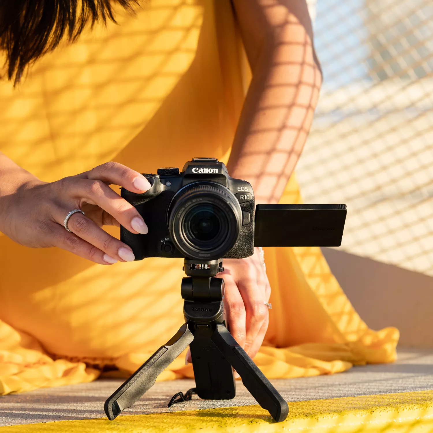 Woman holding a Canon camera on a tripod. She is wearing a yellow dress and rings.