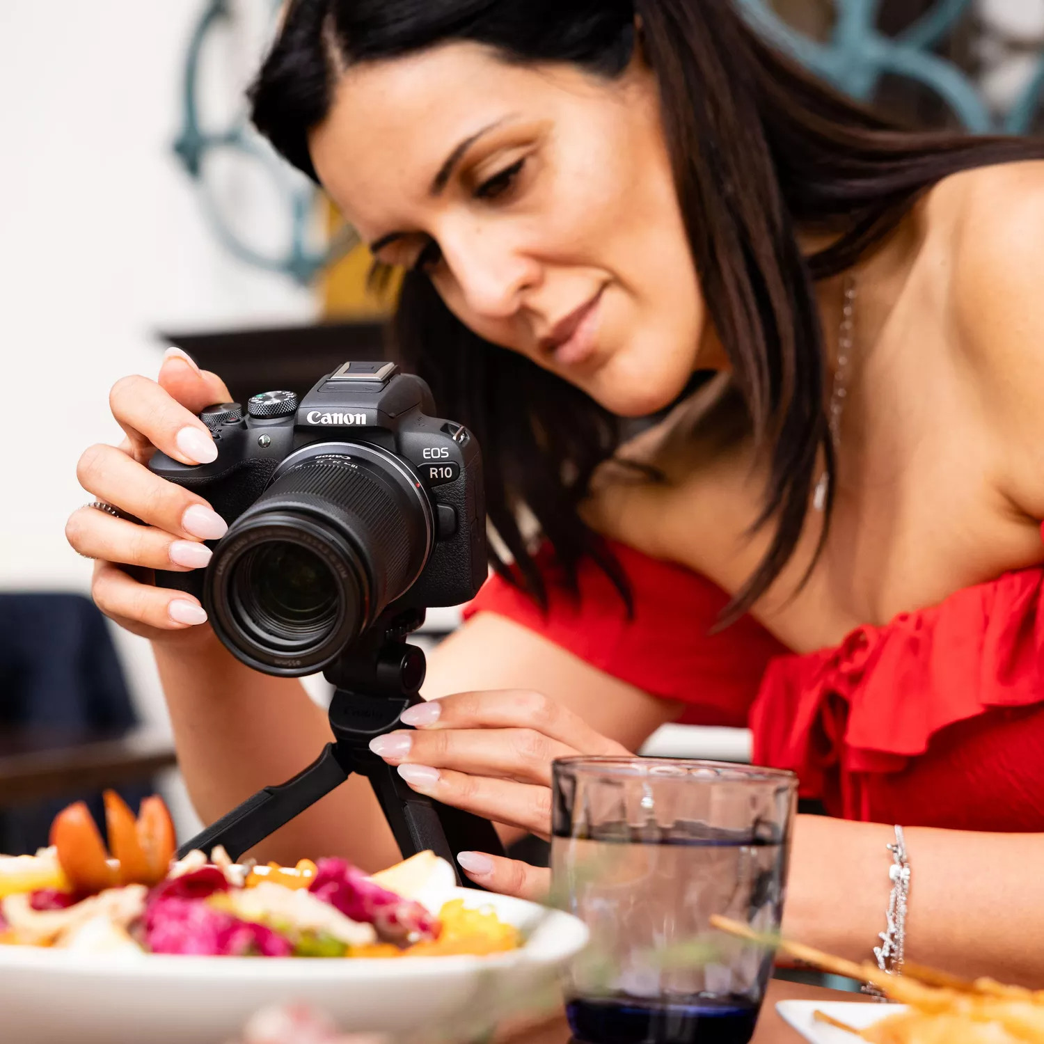 Woman holding a Canon EOS R10 camera, taking photos of food, with a glass of water nearby.