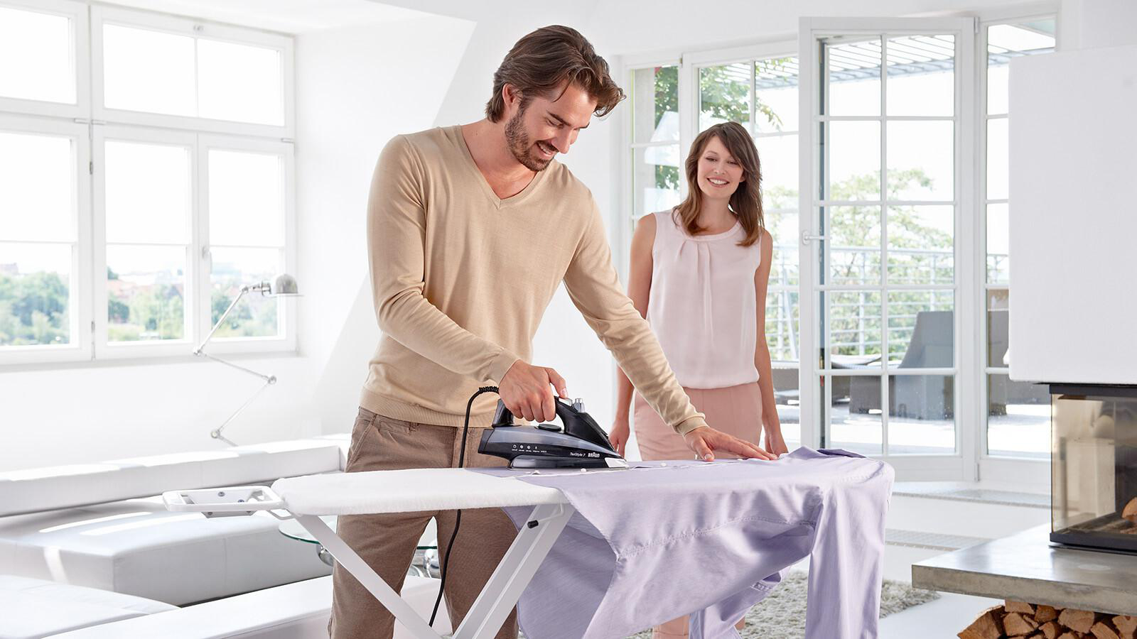 Ein Mann bügelt ein Hemd, während eine Frau zusieht. Helles, modernes Wohnzimmer.