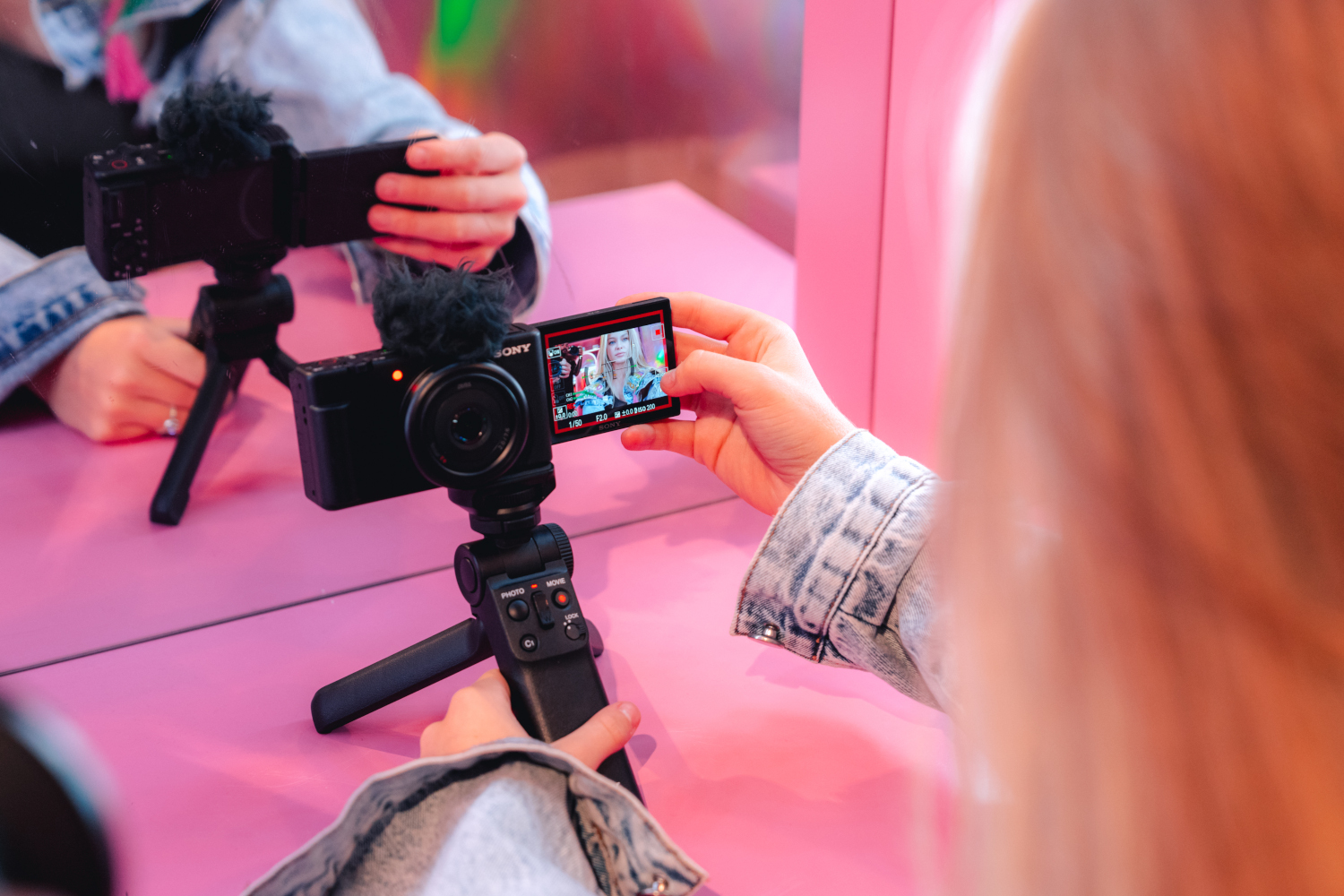 Personne filmant avec une caméra sur trépied, reflétée dans un miroir, fond rose.