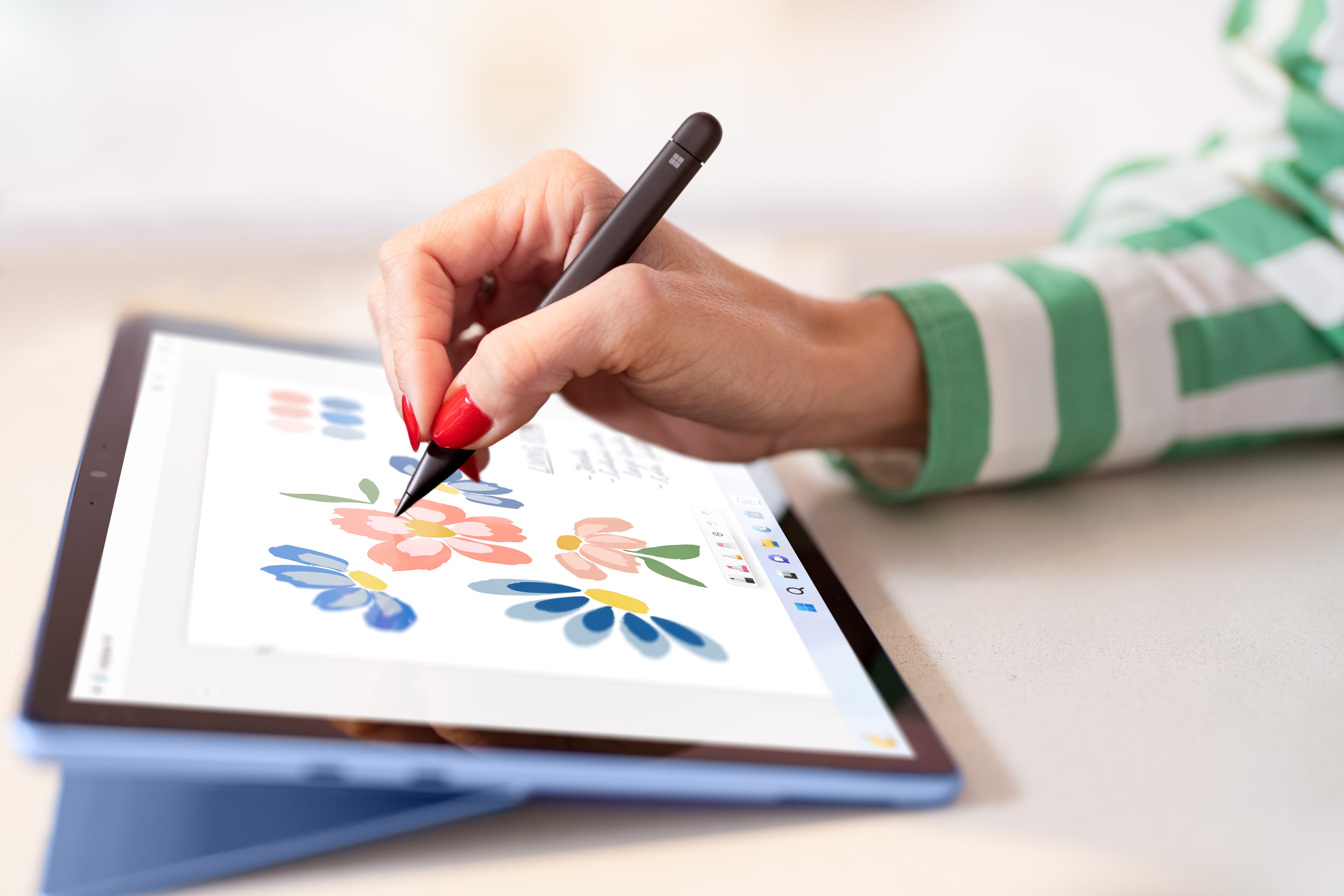 Een hand met rode nagels gebruikt een stylus om bloemen op een tablet te tekenen. Een groen-witte gestreepte mouw is rechts.