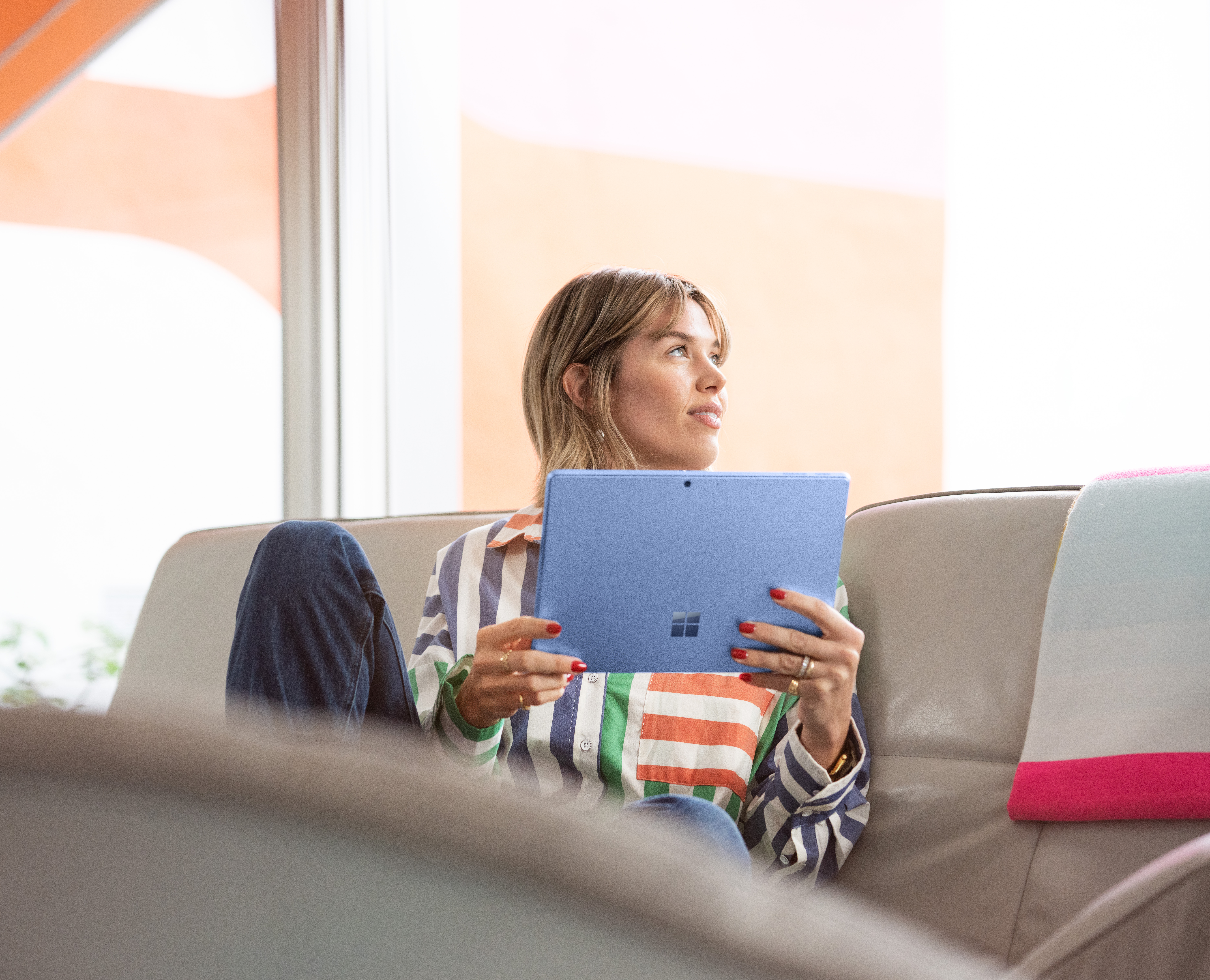 Een vrouw zit op een bank met een blauwe tablet, gekleed in een gestreept shirt. Ze kijkt omhoog, met een raam en een oranje muur op de achtergrond.