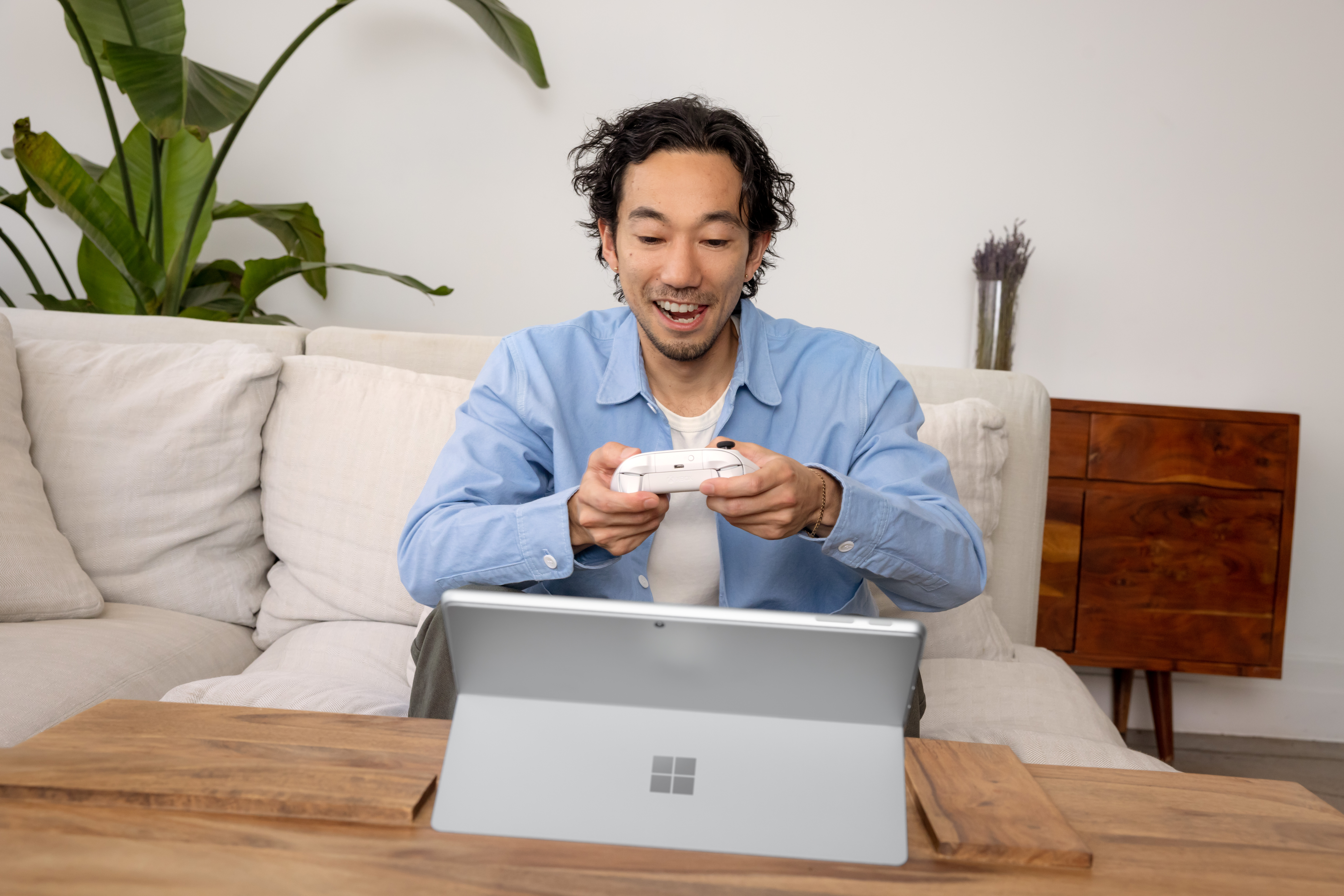Een man speelt een spel, houdt een witte controller vast en kijkt naar een laptop. Een plant en een bank staan op de achtergrond.