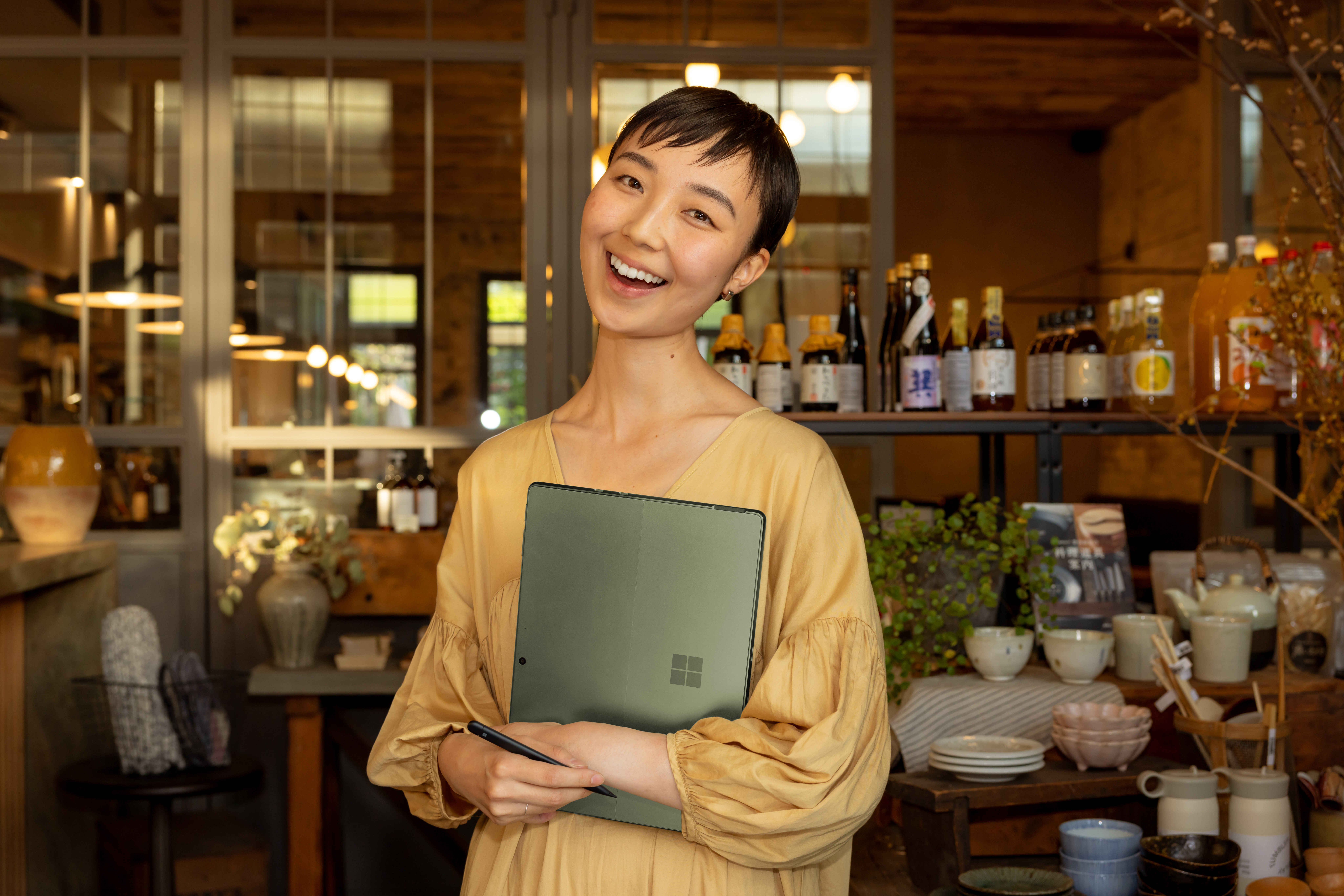 Een vrouw glimlacht, met een laptop en pen. Ze is in een café, met schappen vol flessen en decoratie op de achtergrond.